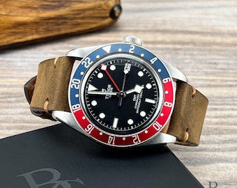 May include: A black dial wristwatch with a red and blue bezel. The watch has a brown leather strap and the words "TUDOR GENEVE GMT CHRONOMETER OFFICIALLY CERTIFIED" printed on the face.