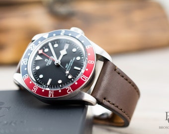 May include: A black-faced wristwatch with a brown leather band. The watch face has a red and blue bezel with white numbers and a date window. The watch face also has the text "TUDOR" and "GMT" printed on it.