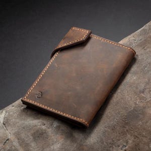 May include: A brown leather wallet with a flap closure, resting on a textured, gray-brown surface. The wallet has visible stitching and a small embossed logo. The background is dark, creating a contrast with the wallet.