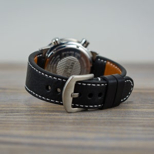 May include: A black leather watch strap with white stitching and a silver buckle. The strap is attached to a silver watch with a black face.