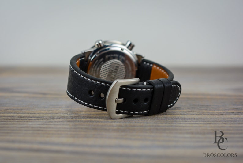 May include: A black leather watch strap with white stitching and a silver buckle. The strap is attached to a silver watch with a black face.