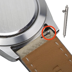 Pode incluir: Close-up de uma pulseira de relógio prateada com uma ferramenta de barra de mola. A ferramenta de barra de mola é usada para remover ou instalar pulseiras de relógio.