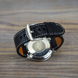 May include: A black leather watch strap with white stitching and a silver buckle. The strap is attached to a silver watch with a black face.