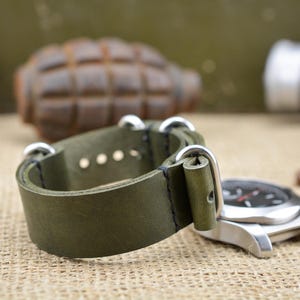 Può includere: Cinturino per orologio in pelle verde oliva con finiture argentate. Il cinturino è abbinato a un quadrante argentato con quadrante nero. Lo sfondo include una granata e proiettili. Il logo "BROSCOLORS" è visibile.