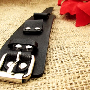 May include: A black leather bracelet with a silver buckle and two silver studs. The bracelet is on a burlap background.