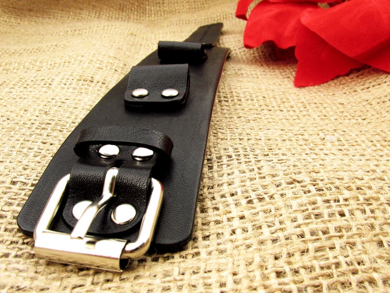 May include: A black leather bracelet with a silver buckle and two silver studs. The bracelet is on a burlap background.