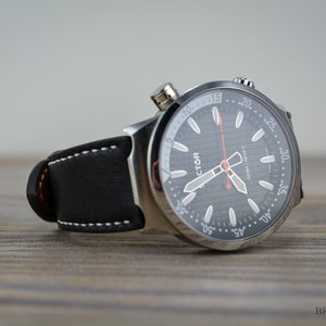 May include: A silver wristwatch with a black face and white markings. The watch has a black leather band with white stitching. The watch face has the word "VICTOR" printed on it.