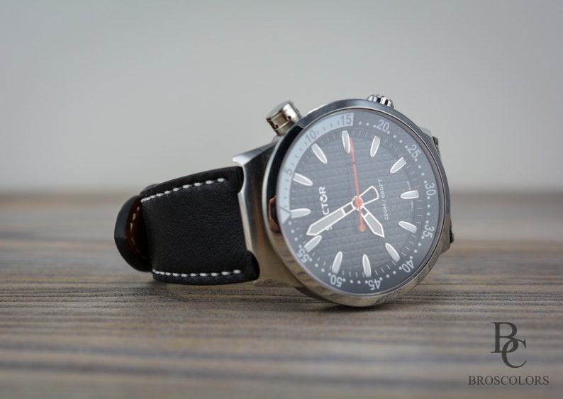 May include: A silver wristwatch with a black face and white markings. The watch has a black leather band with white stitching. The watch face has the word "VICTOR" printed on it.