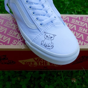 Könnte beinhalten: Weiße Vans-Sneaker aus Canvas mit einer schwarzen Linienzeichnung einer Katze auf der Zehenkappe. Die Schuhe stehen auf einem Vans-Schuhkarton mit dem Vans-Logo, das auf dem Karton wiederholt wird.