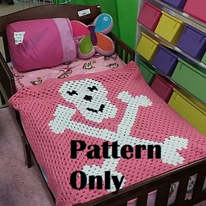 May include: A pink crocheted blanket with a white skull and crossbones design is displayed on a small wooden bed. The words "Pattern Only" are printed on the blanket. A pink pillow and a butterfly-shaped pillow are also on the bed.