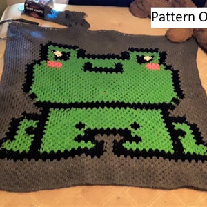 May include: A crocheted blanket with a green frog design. The frog is outlined in black and has pink cheeks. The blanket is gray with a black border.