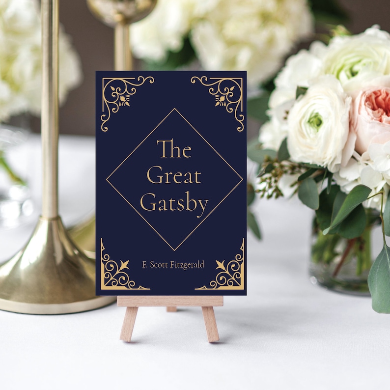 May include: A navy blue card with gold lettering and decorative borders. The card reads "The Great Gatsby" by F. Scott Fitzgerald. The card is displayed on a small wooden easel.