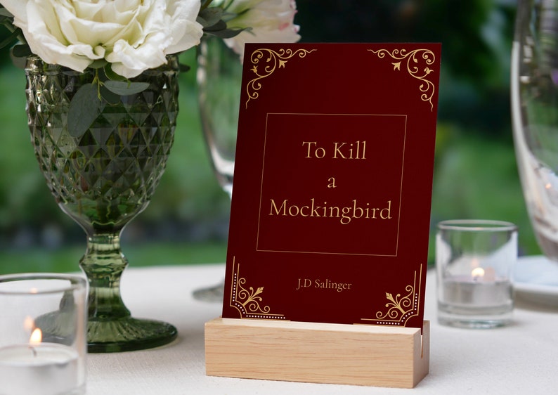 May include: A red book with gold lettering on the cover. The title is "To Kill a Mockingbird" by J.D. Salinger. The book is on a wooden stand.