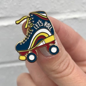 May include: A gold-tone roller skate pin with a blue, red, and yellow design. The skate has white wheels and the text "Let's Roll" is written on the side.