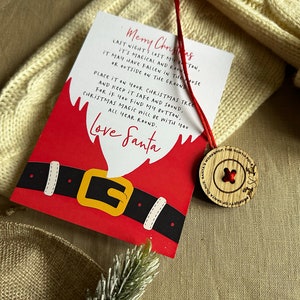 May include: A red and white Christmas card featuring a cartoon Santa Claus. The card includes a message that reads "Merry Christmas. Last night I lost my button, it's magical and rare. It may have fallen in the house or outside on the ground. Place it on your Christmas tree and keep it safe and sound. For if you find my button, Christmas magic will be with you all year round. Love Santa". A wooden button with a red button is attached to the card with a red string.