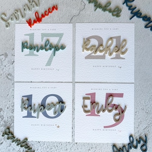 May include: Four white cards with colorful numbers and names on them. The cards say "Wishing you a very" followed by the number and name. The names are Penelope, Rachel, Rupert, and Emily. The numbers are 17, 21, 10, and 15. The cards are surrounded by wooden letters with the names Sarah, Rebecca, Campbell, Kuper, Stanley, and Andy.