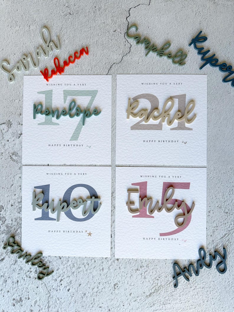 May include: Four white cards with colorful numbers and names on them. The cards say "Wishing you a very" followed by the number and name. The names are Penelope, Rachel, Rupert, and Emily. The numbers are 17, 21, 10, and 15. The cards are surrounded by wooden letters with the names Sarah, Rebecca, Campbell, Kuper, Stanley, and Andy.