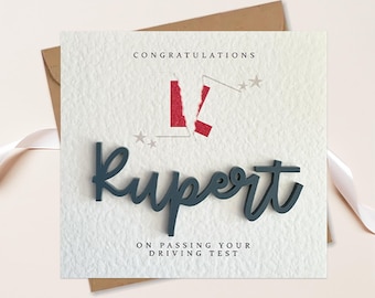 Op de afbeelding: Een witte kaart met de tekst "Gefeliciteerd Rupert met het behalen van je rijbewijs." Een rood-witte L-plaat is doormidden gescheurd en een blauwe 3D-naam "Rupert" staat op de kaart.