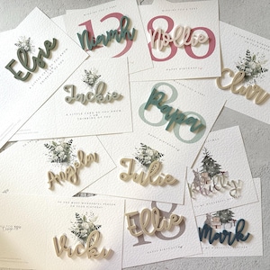 May include: A collection of personalized wooden name cards in various colors, including green, gold, and pink. The cards are decorated with floral designs and feature names such as Ellie, Jackie, Angela, Julie, Ellie, and Mark.