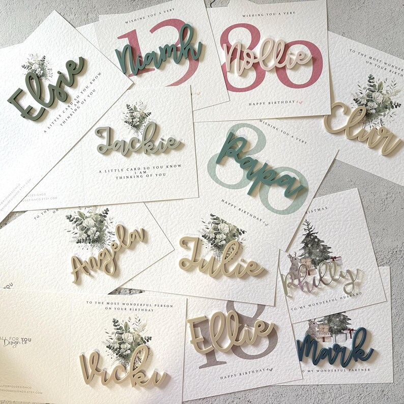May include: A collection of personalized wooden name cards in various colors, including green, gold, and pink. The cards are decorated with floral designs and feature names such as Ellie, Jackie, Angela, Julie, Ellie, and Mark.