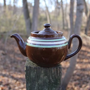 May include: A brown teapot with a white and green striped design sits on a wooden post. The teapot has a lid and a handle.