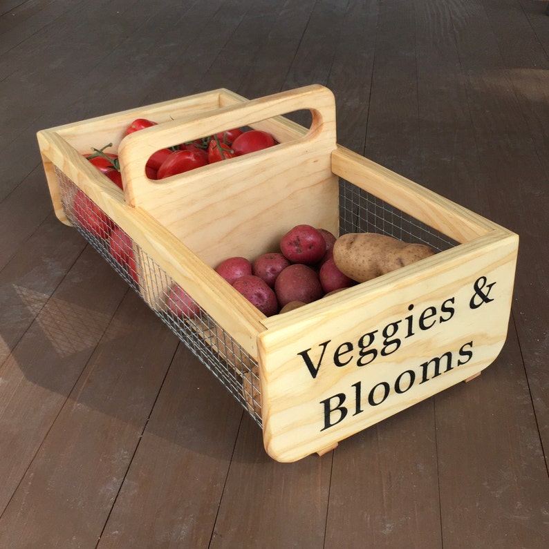 Op de afbeelding: Een houten kist met een draadgaas inzet en een handvat. De kist is gelabeld met "Veggies & Blooms". De kist is gevuld met rode tomaten en rode aardappelen.