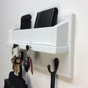May include: White wooden wall mounted key holder with a shelf for mail or other items. It has two hooks and a small shelf. There are keys, a wallet, and a phone on the holder.