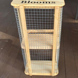 Op de afbeelding: Een houten standaard met draadgaas zijkanten en planken. De standaard heeft een houten bovenkant met de woorden "Veggies & Blooms" erop geschreven.