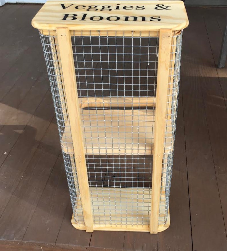 Op de afbeelding: Een houten standaard met draadgaas zijkanten en planken. De standaard heeft een houten bovenkant met de woorden "Veggies & Blooms" erop geschreven.