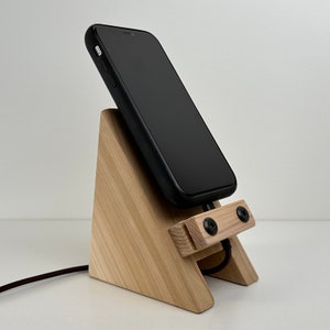 May include: A wooden phone stand with a black smartphone resting on top. The stand has two small black screws on the front and a black cable is connected to the phone.