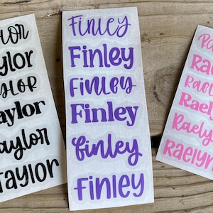 May include: Three sheets of name stickers. One sheet has the name "Taylor" in black, another has "Finley" in purple, and the last has "Raelynn" in pink. The names are in a handwritten style font.