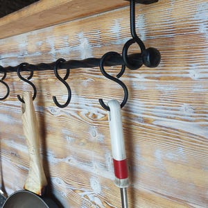 Könnte beinhalten: Schwarzer Schmiedeeisen-Topfhalter mit sechs S-förmigen Haken, montiert an einer Holzwand. Der Halter hält einen Schaumlöffel, einen Holzspatel und ein Küchenutensil mit rot-weißem Griff. Das Holz hat eine helle, rustikale Oberfläche.