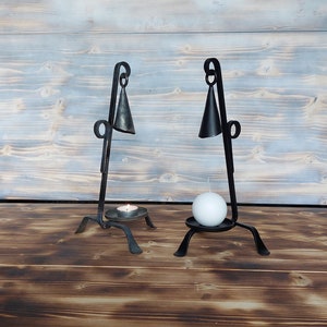May include: Two black metal candle holders with a bell-shaped design. Each holder has a round base and three legs. One holder has a small white candle and the other has a white pillar candle.