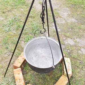 Può includere: Un cavalletto in metallo nero con una catena e un gancio per appendere una pentola sopra un fuoco. Il cavalletto è installato su un letto di mattoni rossi.