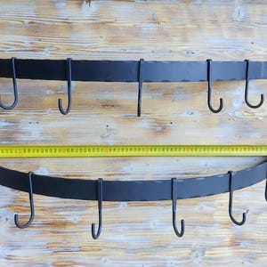 Könnte beinhalten: Zwei schwarze Topfaufhängungen aus Metall mit dekorativen Blumenakzenten und mehreren Haken. Die Aufhängungen sind an einer hellholzfarbenen Oberfläche montiert.