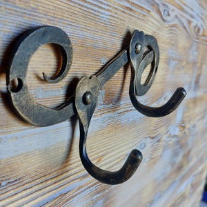 Può includere: Ganci da parete in metallo forgiati a mano con finitura rustica e antica. I ganci sono montati su una superficie in legno con venature naturali. Il design presenta volute e curve decorative.