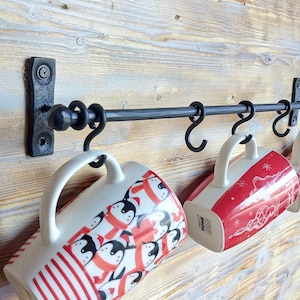 Puede incluir: Un colgador de tazas de metal negro con cuatro ganchos montado en una pared de madera. Cuatro tazas con patrones rojos y blancos cuelgan de los ganchos.