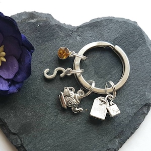 May include: A silver charm keychain with a teapot, tea bag, and letter S charm. The keychain is on a gray heart-shaped stone.