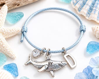 Könnte beinhalten: Ein hellblaues Kordelarmband mit drei silberfarbenen Anhängern: einem detaillierten Hai, einem runden, klaren Strassstein und dem Buchstaben D. Das Armband verfügt über verstellbare Schiebeknoten auf beiden Seiten, um den Sitz anzupassen.