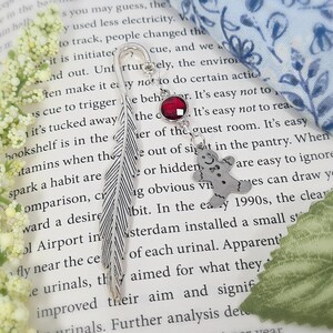 May include: A silver feather bookmark with a red rhinestone and a silver gingerbread man charm. The bookmark is on a page of a book with white flowers in the background.