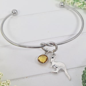 Peut inclure: Un bracelet jonc argenté avec un nœud, orné d'un petit charme rond en pierre jaune et d'un charm en forme de kangourou. Le bracelet est présenté sur une surface blanche avec des éléments floraux.
