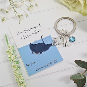 Könnte beinhalten: Eine blaue und weiße Karte mit einer Rochen-Illustration und dem Text "Your Personalised Message Here" und "Love from... Whispering Words Gifts". Ein silberner Schlüsselanhänger mit einem Rochen-Anhänger, einem Buchstaben M-Anhänger und einem blauen Kristall-Anhänger.