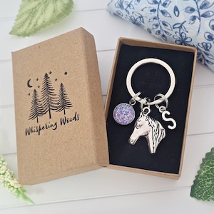 May include: A silver horse head keyring with a purple glitter charm and a silver charm with the letter "S". The keyring is in a brown box with the words "Whispering Woods" printed on it.
