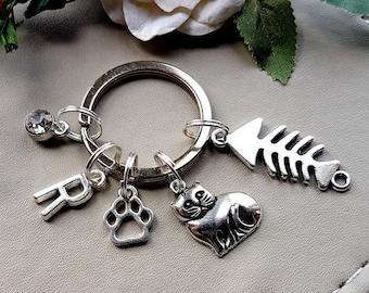May include: Silver keychain with a ring and multiple charms. Charms include a letter "R", a paw print, a cat, a fishbone, and a crystal. The keychain is on a light gray surface with floral accents.