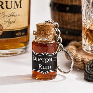 Könnte beinhalten: Ein kleiner Schlüsselanhänger in Form einer Glasflasche, gefüllt mit bernsteinfarbener Flüssigkeit, mit einem Korkstopfen und einem schwarzen Etikett mit weißem Text Emergency Rum. Die Flasche ist an einer silberfarbenen Kette und einem Ring befestigt.