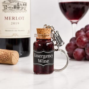 May include: A miniature glass bottle keyring filled with dark red liquid, topped with a cork stopper. A black label on the front features the white text Emergency Wine. A silver-toned metal charm shaped like a corkscrew is attached to the keyring.