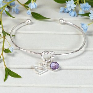 May include: A silver-toned bangle bracelet with a knot design in the center. It features a charm with the number 27 and a round, purple gemstone. The bracelet has a simple, elegant design.