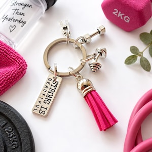 May include: A silver-toned metal keychain featuring a rectangular charm engraved with the text STRONG IS BEAUTIFUL, a miniature dumbbell charm, and a bright pink suede-style tassel, all attached to a circular split ring.