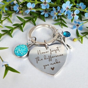 May include: Silver keyring with a heart-shaped charm engraved with the words "Never forget the difference you make." The keyring also features a round, turquoise charm and a blue crystal accent. The background includes blue flowers and green leaves.