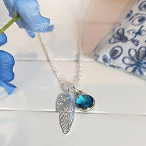 May include: A silver necklace with a delicate wing pendant and a round, teal-colored gemstone. The necklace is displayed on a white surface, with a blue flower and patterned fabric in the background. The necklace is a piece of jewelry.
